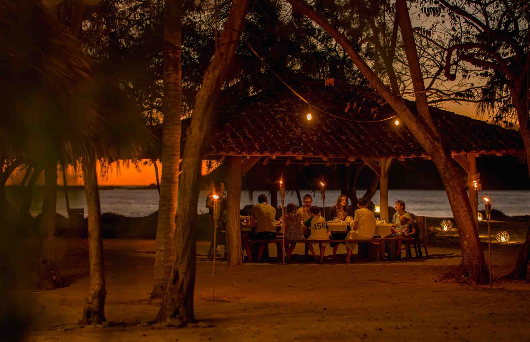 Families share dinner under string lights by the beach at dusk